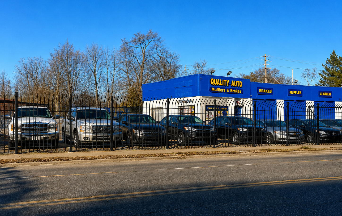 Quality Auto Mufflers & Brakes on Fenkell Avenue in Detroit, blue and yellow storefront with brake, muffler and alignment signs, customer cars parked in the fenced lot.